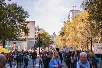 A large group of people are walking down a tree-lined city street, participating in a protest or demonstration. Some hold banners and signs, and there is a sense of movement and engagement among the diverse crowd. City buildings and construction cranes are visible in the background, suggesting an urban setting.