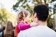 A young child with curly hair is held by an adult figure, likely a caregiver, amidst a blurred outdoor background with green foliage. The child wears a pink long-sleeve shirt and an orange vest, clutching a yellow toy or cloth.