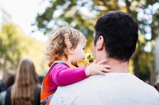 A young child with curly hair is held by an adult figure, likely a caregiver, amidst a blurred outdoor background with green foliage. The child wears a pink long-sleeve shirt and an orange vest, clutching a yellow toy or cloth.