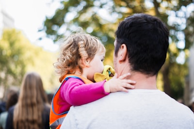 A young child with curly hair is held by an adult figure, likely a caregiver, amidst a blurred outdoor background with green foliage. The child wears a pink long-sleeve shirt and an orange vest, clutching a yellow toy or cloth.
