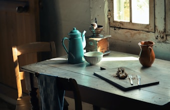 A rustic kitchen scene with a wooden table set near an old window. On the table are a vintage coffee pot, a white bowl, a brown ceramic jug, and a wooden coffee grinder. A bread roll with crumbs is placed on a cutting board with two butter knives. Light filters softly through the window, casting gentle shadows and highlighting the aged textures of the furniture.