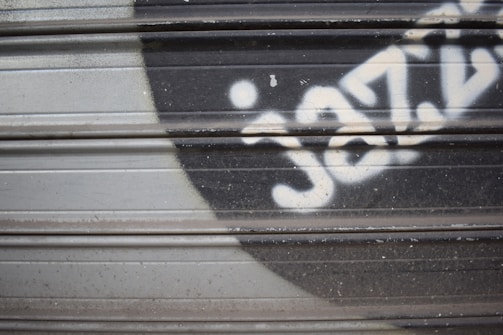 Metal shutters with graffiti featuring stylized text. A prominent black and white design is visible against the corrugated metal surface.