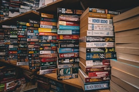 Stacks of paperback books are arranged on wooden shelves in a somewhat cluttered manner. Various genres and titles are visible, featuring works by authors such as John Sandford and W.E.B. Griffin. The scene conveys a sense of an extensive, varied collection typical of a well-stocked bookstore or personal library.