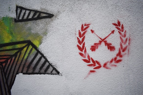 A wall features a graffiti design on the left with geometric shapes in black, yellow, and green spray paint. On the right, there is a red stencil design of laurel wreaths encircling two crossed arrows.