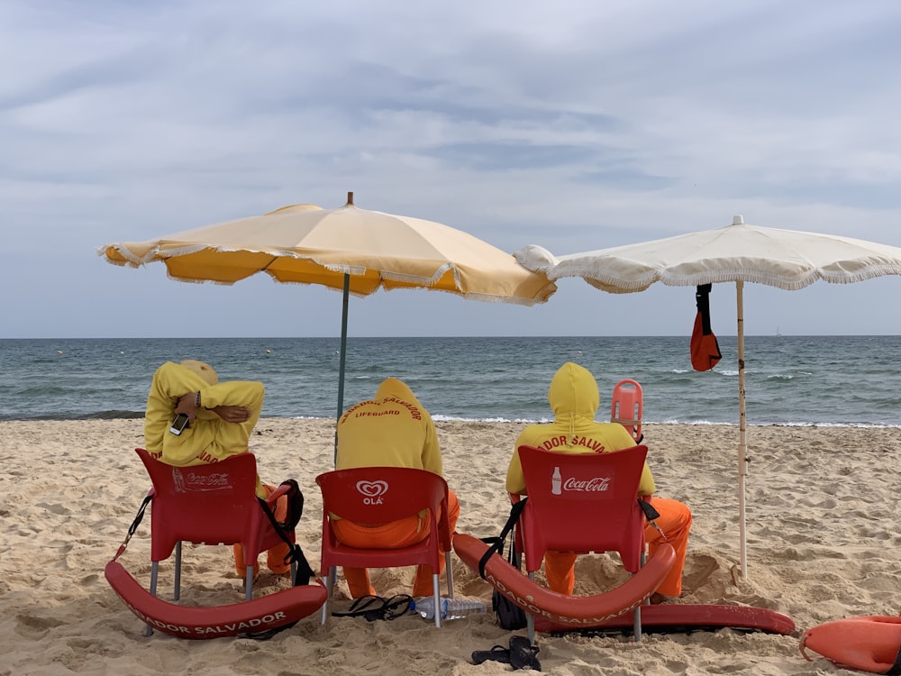 three persons sitting on chairs under parasols at the sand seashore during day