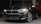 A sleek black BMW car is parked with its headlights on, the grille prominently displayed. The setting appears to be a dimly lit parking garage or industrial space.