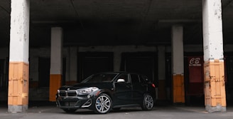 A sleek dark green car parked in front of a modern warehouse with shipping containers.