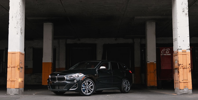A sleek dark green car parked in front of a modern warehouse with shipping containers in the background.