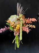 A bouquet composed of various flowers and leaves, artistically arranged. The flowers display a mix of textures and colors, including green hydrangeas, orange and brown leaves, and spiky tall grasses. The arrangement is held against a dark background that enhances the vivid colors of the bouquet.
