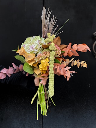A bouquet composed of various flowers and leaves, artistically arranged. The flowers display a mix of textures and colors, including green hydrangeas, orange and brown leaves, and spiky tall grasses. The arrangement is held against a dark background that enhances the vivid colors of the bouquet.