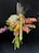 A bouquet composed of various flowers and leaves, artistically arranged. The flowers display a mix of textures and colors, including green hydrangeas, orange and brown leaves, and spiky tall grasses. The arrangement is held against a dark background that enhances the vivid colors of the bouquet.