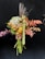 A bouquet composed of various flowers and leaves, artistically arranged. The flowers display a mix of textures and colors, including green hydrangeas, orange and brown leaves, and spiky tall grasses. The arrangement is held against a dark background that enhances the vivid colors of the bouquet.