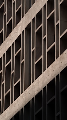 A geometric facade of a building with a series of parallel and perpendicular lines creating a grid-like structure. The concrete surface features a prominent diagonal element that adds visual interest through its contrasting angle.