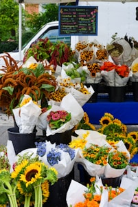 Bouquets of various colorful flowers are arranged in black buckets, wrapped in white paper, and displayed for sale at a market stall. A handwritten sign lists the prices and types of flowers. The scene is vibrant and lively, with sunflowers, marigolds, and other blooms creating a diverse and appealing display.
