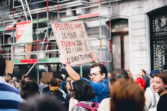 A crowd of people gathered in a protest on the street, with one person prominently holding a handmade sign that reads, 'Politicians do your fucking job so I can go back to mine.' The background shows a building with scaffoldings and various other signs.