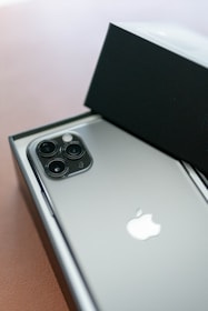 A smartphone with a sleek metallic back and triple camera setup is partially inside an open box. The Apple logo is prominently visible on the back of the phone.