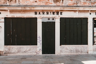 The image depicts an old, closed shop front with a weathered façade, displaying the word 'BARBIERE' above dark, boarded windows and door. The building shows signs of age with worn brickwork and faded paint. There is graffiti on the wall to the right.