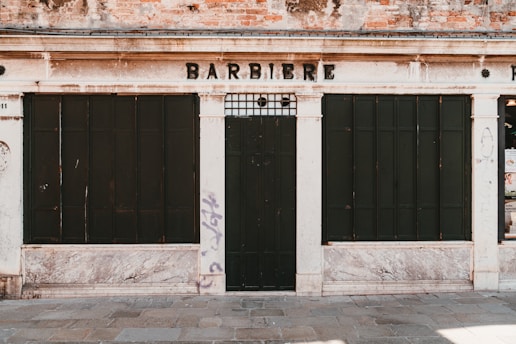 The image depicts an old, closed shop front with a weathered façade, displaying the word 'BARBIERE' above dark, boarded windows and door. The building shows signs of age with worn brickwork and faded paint. There is graffiti on the wall to the right.