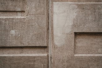 A textured concrete wall featuring rectangular patterns and subtle variations in color. The surface appears worn and slightly uneven with visible lines and angular shapes.