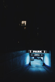 A dimly lit entrance to an underground parking garage, with glowing blue lights illuminating the walls and ground. A sign above reads 'PARK' in bold letters. A single parked car is visible inside. The scene is mostly dark, with a lighted window visible in the upper background of a building.