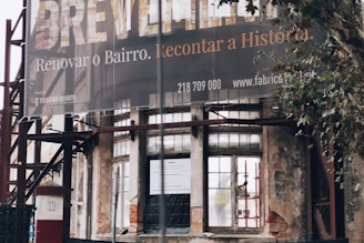 An exterior view of a partially renovated building with an old, weathered facade. A large banner hangs above with a message in Portuguese about renovating the neighborhood. The building's walls show signs of decay, with visible scaffolding and an urban setting surrounding it. A tree is seen to the right of the image.