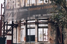 An exterior view of a partially renovated building with an old, weathered facade. A large banner hangs above with a message in Portuguese about renovating the neighborhood. The building's walls show signs of decay, with visible scaffolding and an urban setting surrounding it. A tree is seen to the right of the image.
