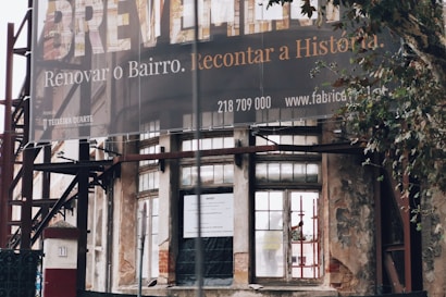 An exterior view of a partially renovated building with an old, weathered facade. A large banner hangs above with a message in Portuguese about renovating the neighborhood. The building's walls show signs of decay, with visible scaffolding and an urban setting surrounding it. A tree is seen to the right of the image.