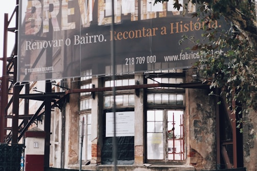 An exterior view of a partially renovated building with an old, weathered facade. A large banner hangs above with a message in Portuguese about renovating the neighborhood. The building's walls show signs of decay, with visible scaffolding and an urban setting surrounding it. A tree is seen to the right of the image.