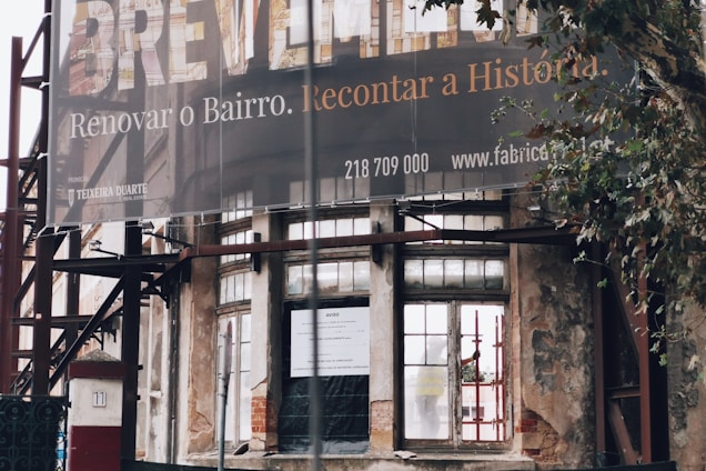 An exterior view of a partially renovated building with an old, weathered facade. A large banner hangs above with a message in Portuguese about renovating the neighborhood. The building's walls show signs of decay, with visible scaffolding and an urban setting surrounding it. A tree is seen to the right of the image.