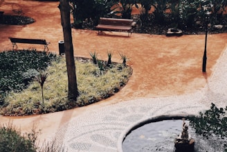 A peaceful park setting featuring a curved path with a mix of tiled and earthy surfaces. Wooden benches are placed along the path, surrounded by a variety of plants and small trees. A small pond or water feature with a statue is visible, adding to the tranquil environment.