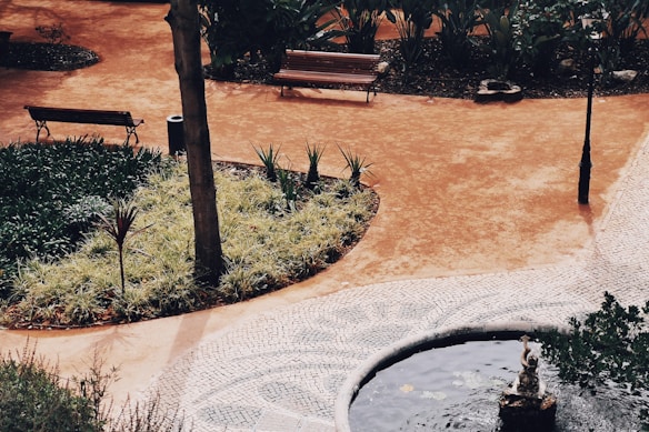 A peaceful park setting featuring a curved path with a mix of tiled and earthy surfaces. Wooden benches are placed along the path, surrounded by a variety of plants and small trees. A small pond or water feature with a statue is visible, adding to the tranquil environment.