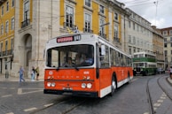 A classic orange and white bus marked 'Reservado' is driving on a cobblestone street lined with colorful historic buildings. Pedestrians are visible on the sidewalk, and a green double-decker bus follows in the background.