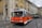 A classic orange and white bus marked 'Reservado' is driving on a cobblestone street lined with colorful historic buildings. Pedestrians are visible on the sidewalk, and a green double-decker bus follows in the background.