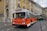 A classic orange and white bus marked 'Reservado' is driving on a cobblestone street lined with colorful historic buildings. Pedestrians are visible on the sidewalk, and a green double-decker bus follows in the background.