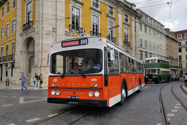 A classic orange and white bus marked 'Reservado' is driving on a cobblestone street lined with colorful historic buildings. Pedestrians are visible on the sidewalk, and a green double-decker bus follows in the background.