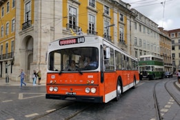 A classic orange and white bus marked 'Reservado' is driving on a cobblestone street lined with colorful historic buildings. Pedestrians are visible on the sidewalk, and a green double-decker bus follows in the background.