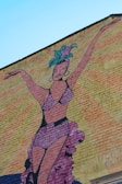 A colorful mural painted on a brick wall depicts a woman in a vibrant costume with her arms raised. She wears a headdress with green and purple elements and a matching purple outfit. The background features rays of orange and yellow, emphasizing a lively and dynamic atmosphere.