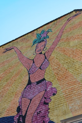 A colorful mural painted on a brick wall depicts a woman in a vibrant costume with her arms raised. She wears a headdress with green and purple elements and a matching purple outfit. The background features rays of orange and yellow, emphasizing a lively and dynamic atmosphere.