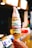 A tall, multicolored soft serve ice cream cone is held up by a hand. The ice cream has layers in various colors including pink, green, blue, and brown, creating a vibrant, eye-catching appearance. The background appears to be indoors, possibly in a café or ice cream shop, with blurred lights and objects.
