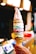 A tall, multicolored soft serve ice cream cone is held up by a hand. The ice cream has layers in various colors including pink, green, blue, and brown, creating a vibrant, eye-catching appearance. The background appears to be indoors, possibly in a café or ice cream shop, with blurred lights and objects.