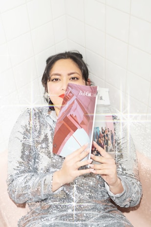 A person wearing a sequined dress leans against a tiled wall and holds a magazine titled 'Bob Cut' covering part of their face. The image has a sparkling, glittery effect overlay.