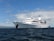 A large white cruise ship named Galapagos Legend is anchored on a calm ocean with a clear blue sky and scattered clouds. The ship features multiple decks and a prominent funnel, indicating it is well-equipped for luxury travel.