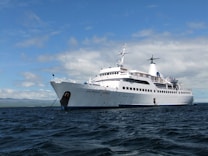 A large white cruise ship named Galapagos Legend is anchored on a calm ocean with a clear blue sky and scattered clouds. The ship features multiple decks and a prominent funnel, indicating it is well-equipped for luxury travel.