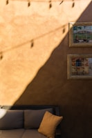 A rustic wall with a warm, earthy tone is partly bathed in sunlight. Two framed artworks depicting outdoor scenes hang on one side. The lower half reveals a cushioned wicker sofa with a single soft pillow. Strings of lights cast shadows across the wall.