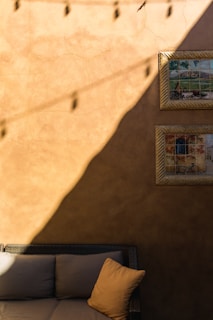 A rustic wall with a warm, earthy tone is partly bathed in sunlight. Two framed artworks depicting outdoor scenes hang on one side. The lower half reveals a cushioned wicker sofa with a single soft pillow. Strings of lights cast shadows across the wall.