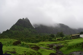 Lush green hillsides rise dramatically against a backdrop of mist-shrouded peaks. The landscape is dotted with vibrant greenery and occasional rocky outcrops. A small structure peeks through the greenery on the right side, adding an element of human presence to the otherwise natural scene.