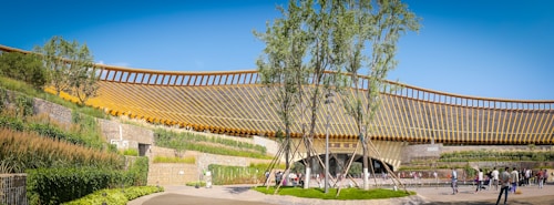 A modern architectural structure with a large, curved design featuring wooden slats forms the central focus. Lush greenery and landscaped walls surround the building, with groups of people walking and gathering in the foreground. The sky is clear and blue, enhancing the vibrancy of the scene.