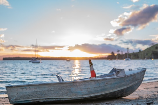 A small boat rests on a sandy shore with a calm body of water extending into the horizon. The sunset creates a warm glow, casting reflections on the water. Several other boats are visible in the distance, softly blurred by the depth of field. The sky is partly cloudy with hues of orange, pink, and blue.