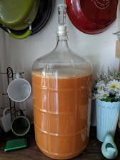 A large glass carboy filled with an orange liquid, possibly juice or wine, is shown on a kitchen countertop. Surrounding it are various kitchen items, including hanging pots and a rack holding ceramic mugs. A tall vase with white and yellow flowers decorates the space alongside a small, bird-shaped watering can.