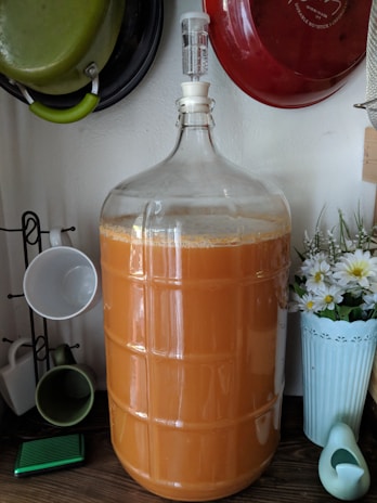 A large glass carboy filled with an orange liquid, possibly juice or wine, is shown on a kitchen countertop. Surrounding it are various kitchen items, including hanging pots and a rack holding ceramic mugs. A tall vase with white and yellow flowers decorates the space alongside a small, bird-shaped watering can.